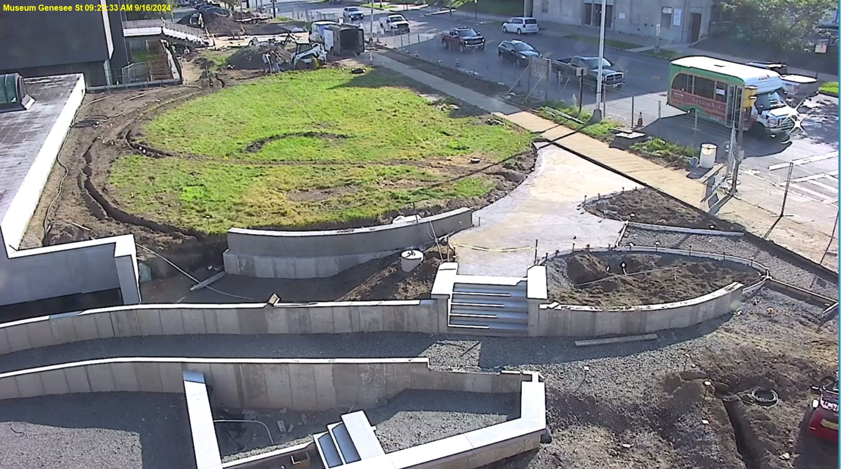 View of Genesee Street front lawn with the museum in the top left corner, the roof of the education wing middle of the left side, street on the right, and showing construction progress in the middle, the concrete sides of the ramp are in progress.