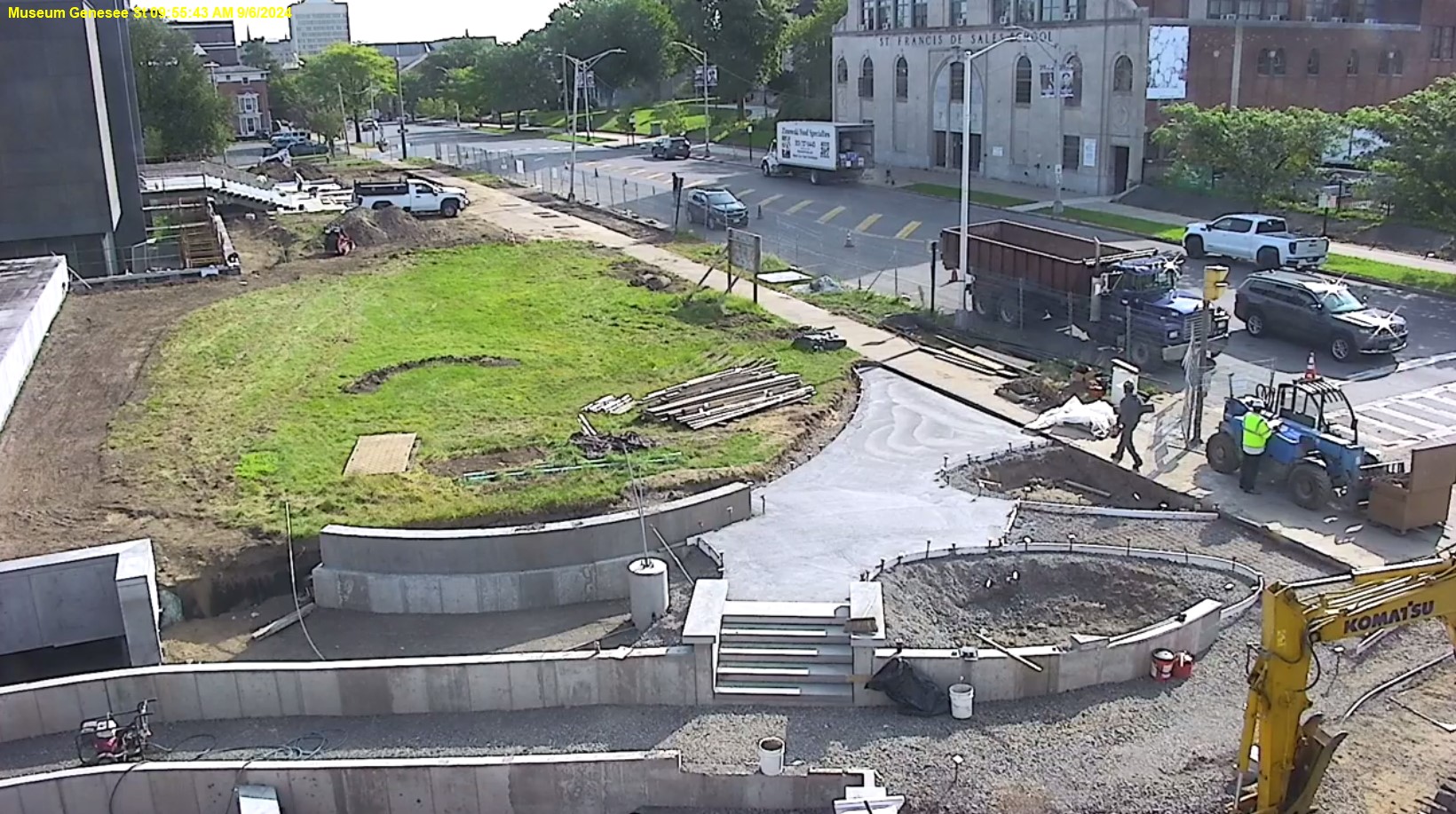 View of Genesee Street front lawn with the museum in the top left corner, the roof of the education wing middle of the left side, street on the right, and showing construction progress in the middle, the concrete sides of the ramp are in progress.